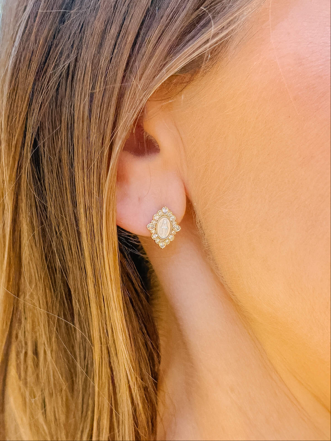 A close-up of a woman's ear wearing a small gold stud earring with a scalloped border and a central oval cream-colored stone.
